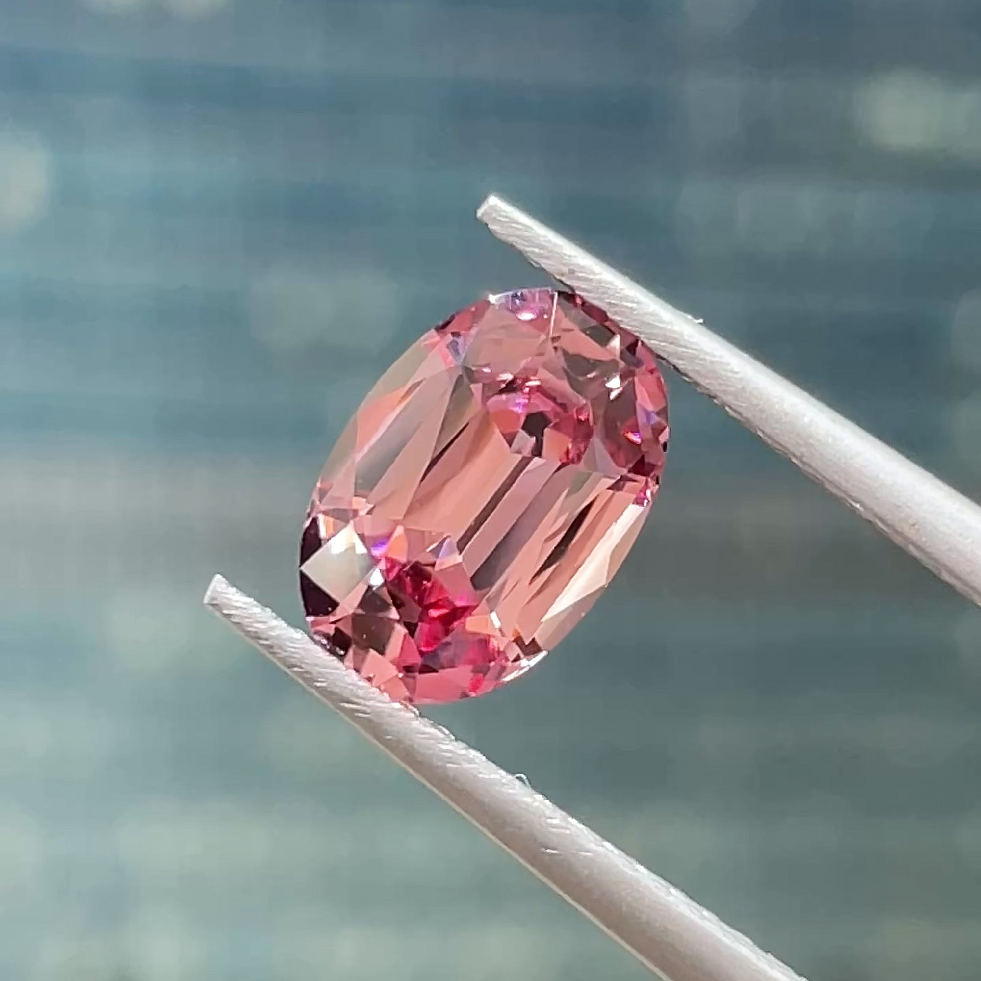 1.74 ct Precision Cut Rose Pink Mahenge Garnet from Tanzania, oval shape, tweezers view on blue grey background